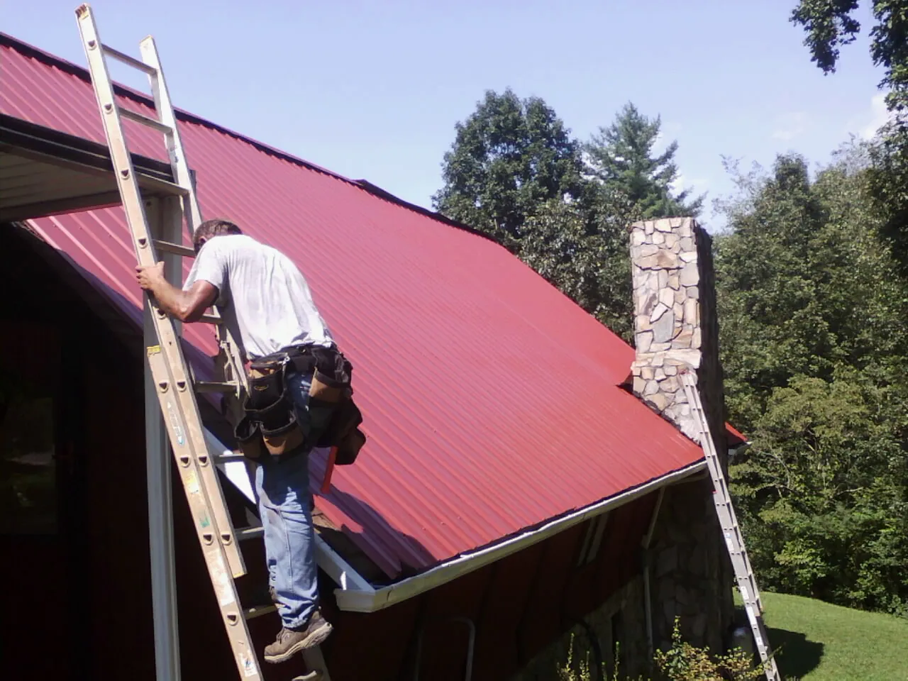Roofer on a ladder beside a red metal roof and stone chimney — Soffit & Fascia Repair by Roofing Brothers in Runnemede